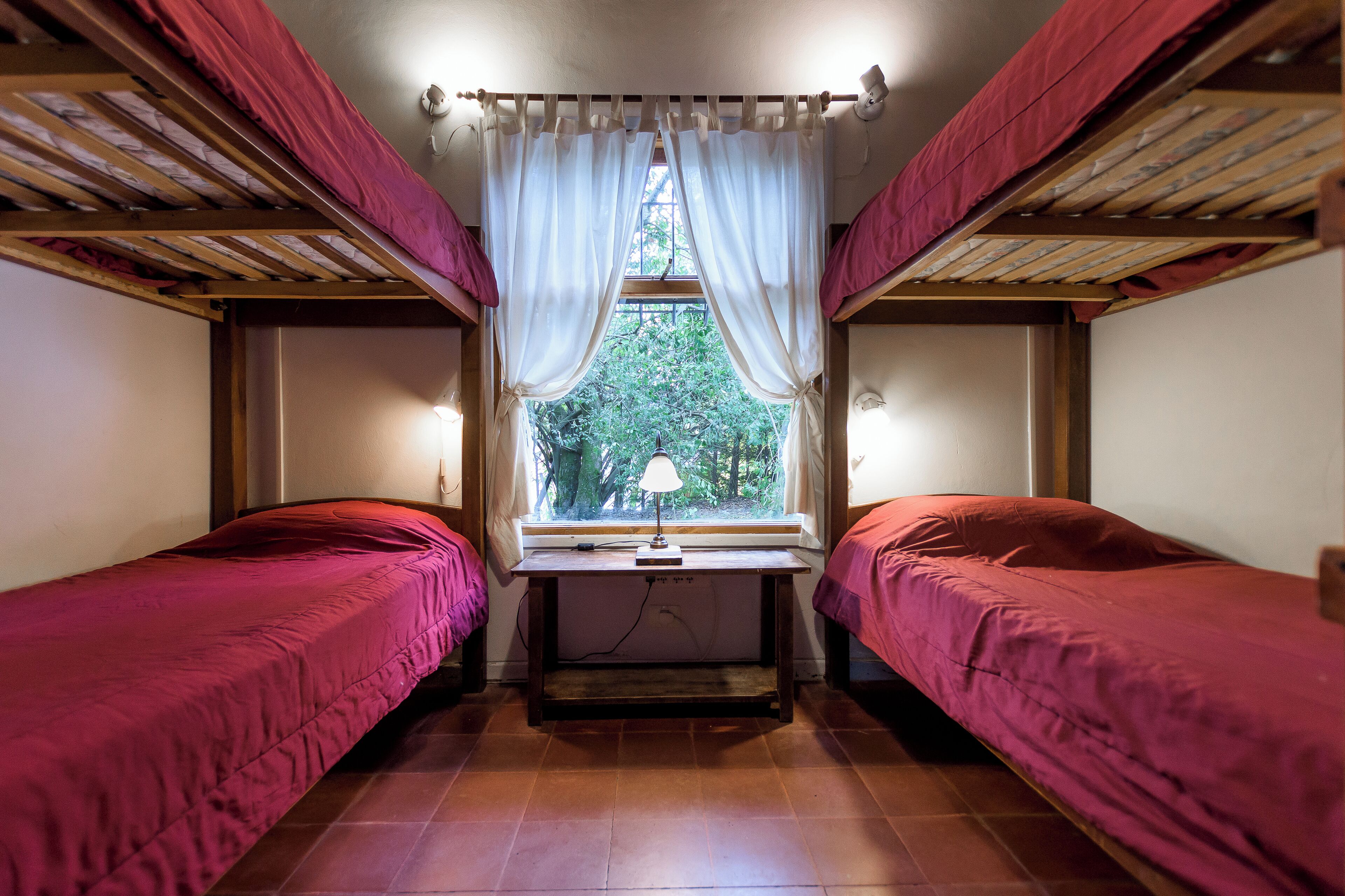 Habitación tipo dormitorio con dos camas, una ventana con cortinas, un escritorio con lámpara y vistas a árboles.