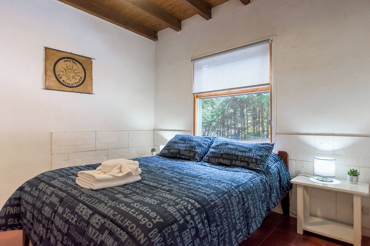 A neatly made bed with a blue patterned bedspread, a small bedside table with a lamp, and a window with a view of trees.