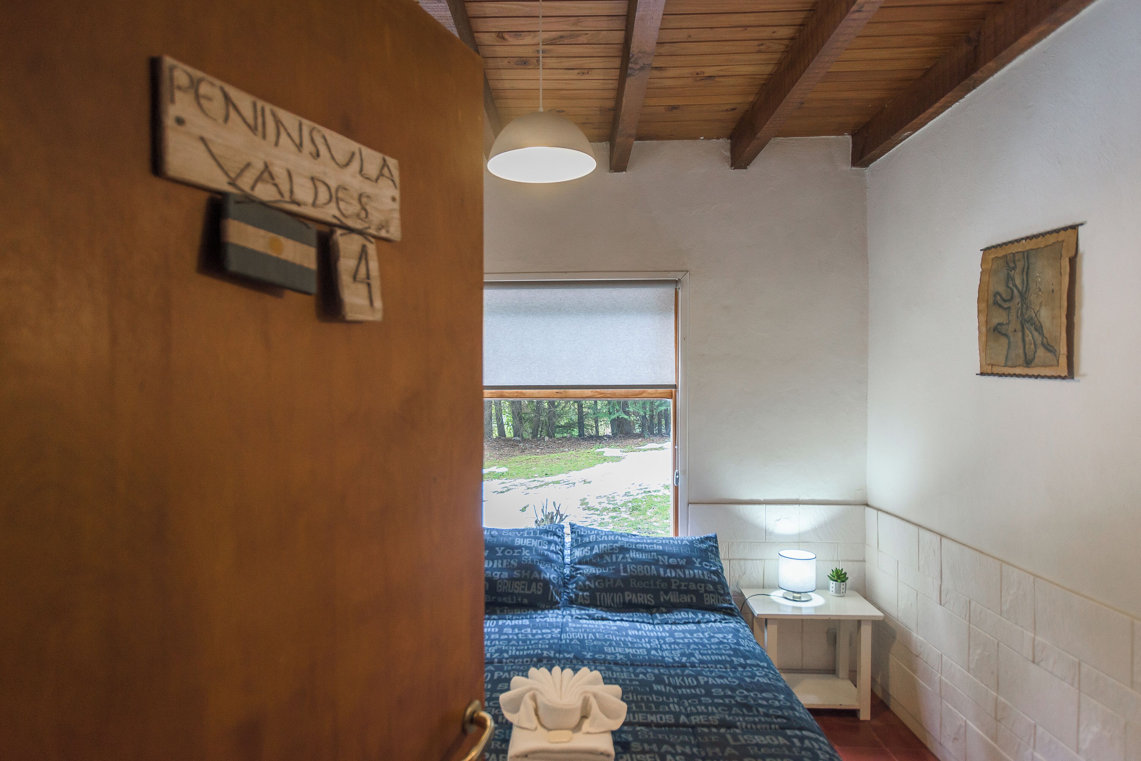 Une chambre avec une porte en bois, un lit recouvert d’une couverture bleue, une table de chevet avec une lampe et une fenêtre donnant sur des arbres.