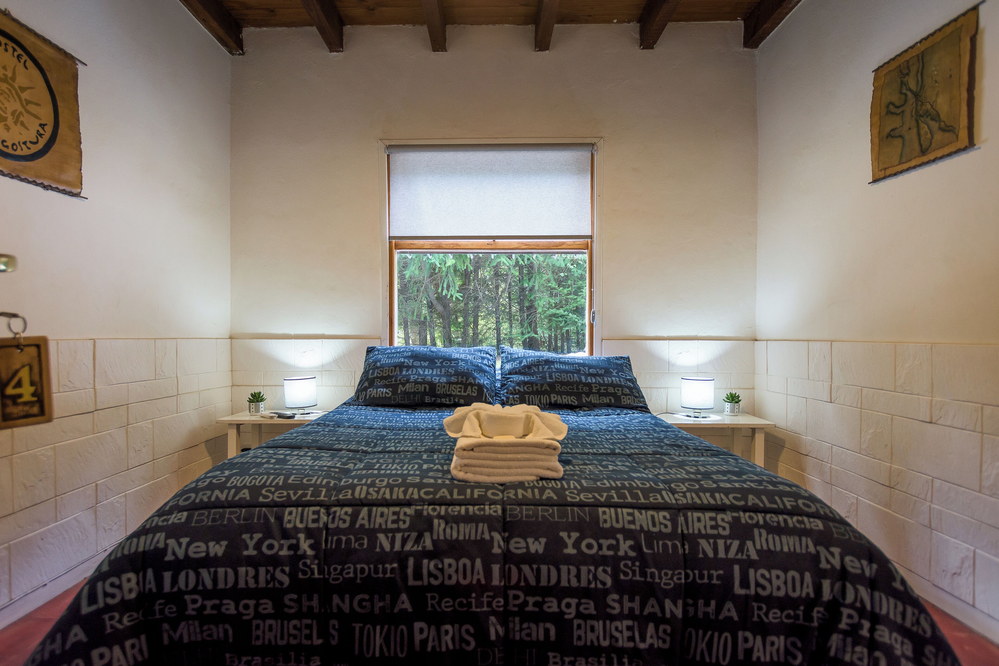 A hotel room with a bed featuring a city-themed bedspread, a window with a view of trees, and a small bedside table with a lamp.