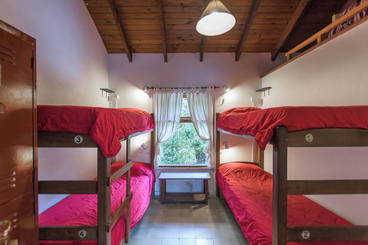 A dormitory room with bunk beds, a window with curtains, and a wooden ceiling.