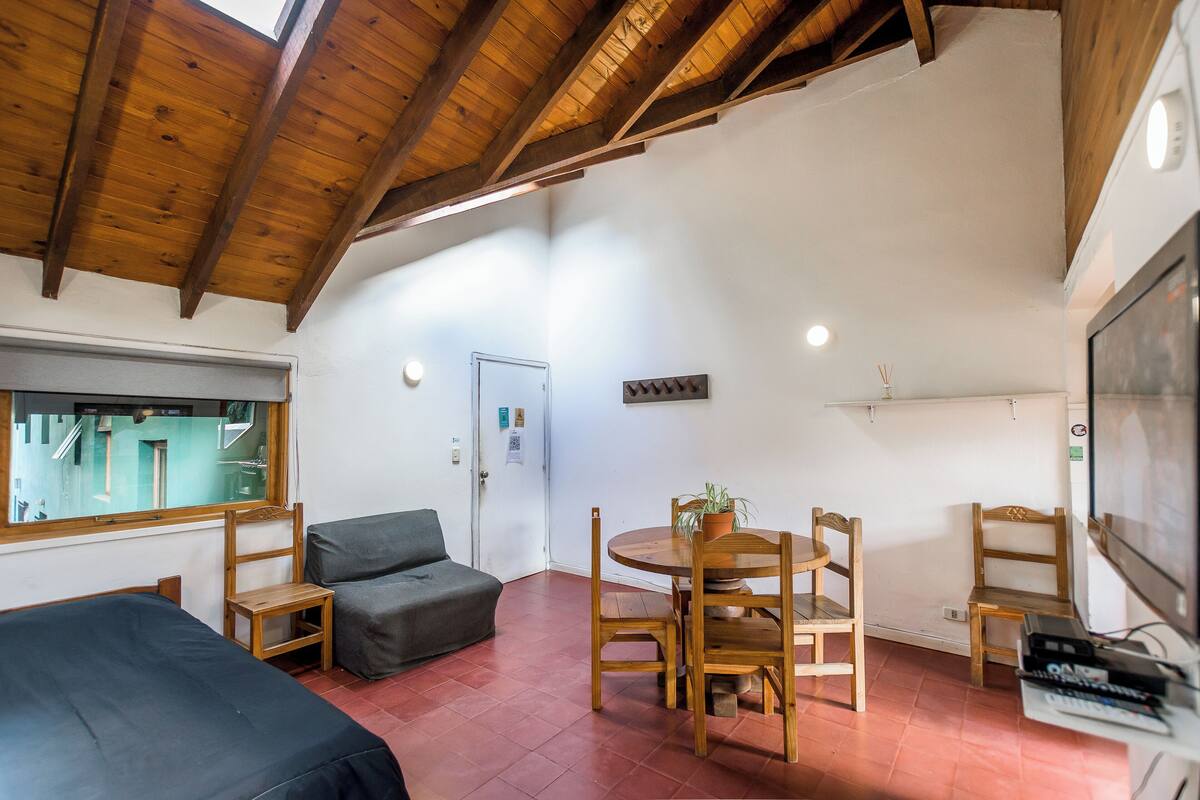 A room with a high wooden ceiling, a dining table with chairs, a sofa, a TV, and a window with a view of buildings.