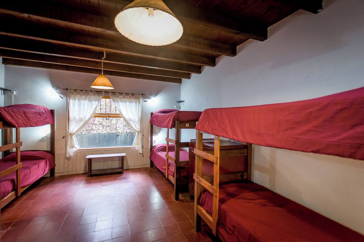 A dormitory room with bunk beds, wooden beams, a window with curtains, and a hanging light fixture.