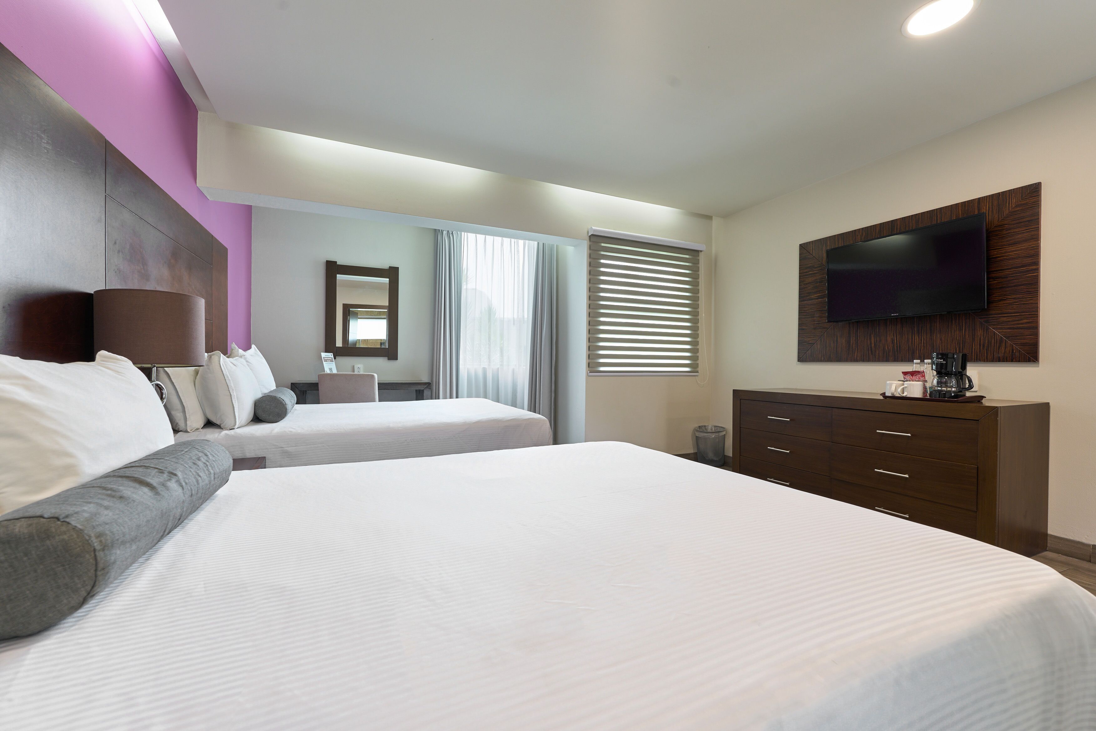 A hotel room with two beds, a TV mounted on the wall, a dresser, and a window with blinds.