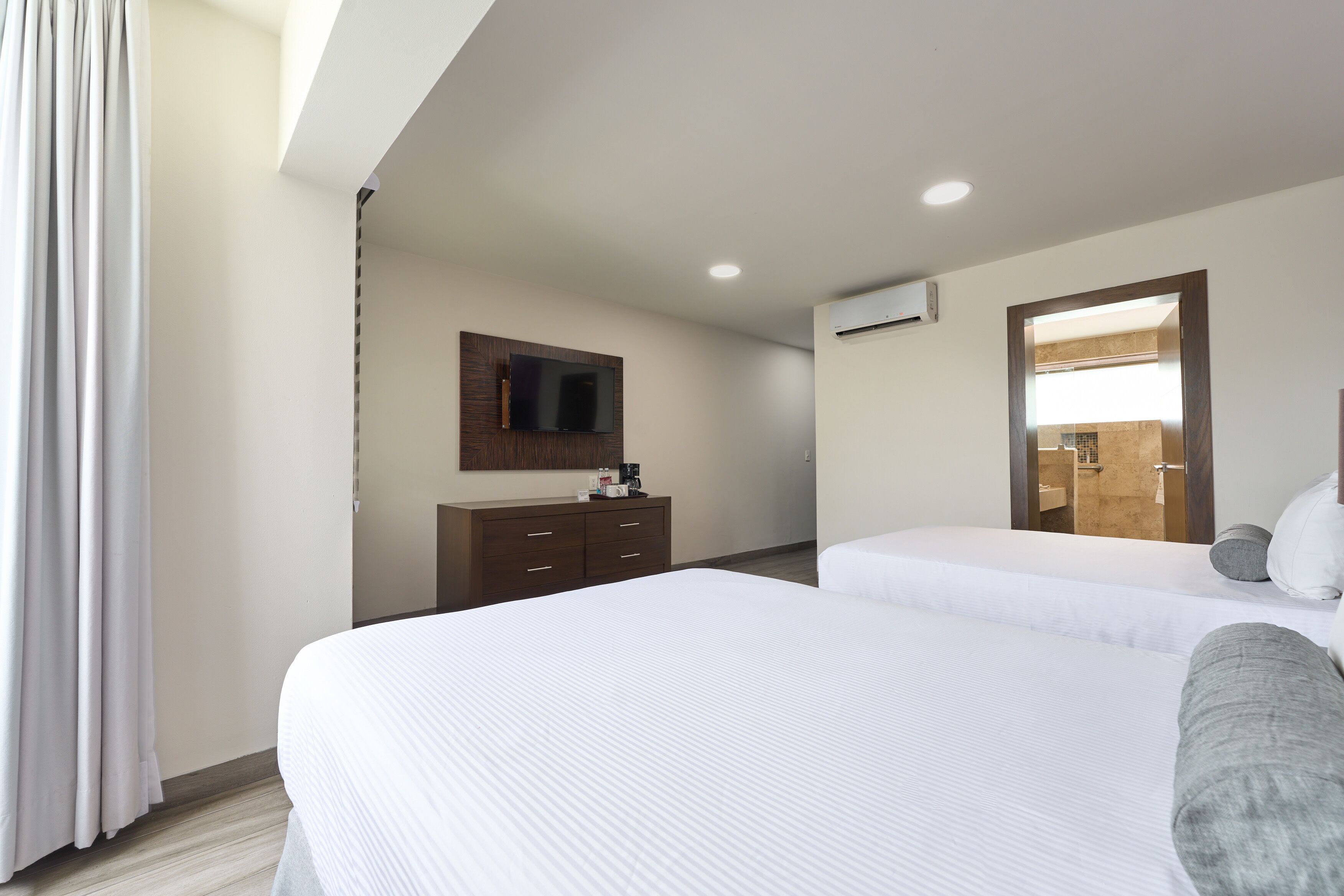 A hotel room with two beds, a wooden dresser, a flat-screen TV, and an air conditioning unit.