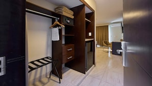 A modern hotel room with a walk-in closet, a desk with a chair, a small refrigerator, and a seating area with a chair and a table. - Hotel Estancia Business Class (Guadalajara)
