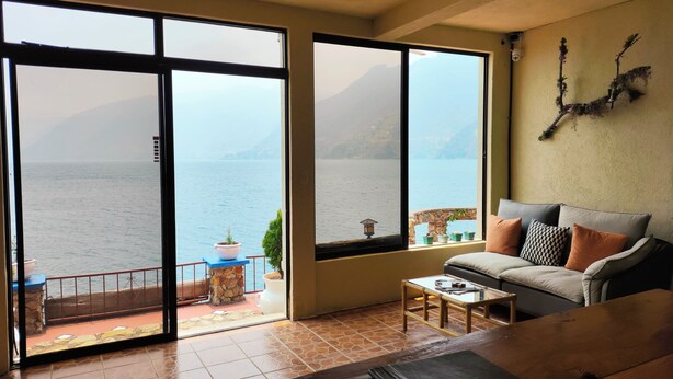 A living room with a large sliding glass door leading to a balcony with a view of the sea and mountains. - Hotel La Villa de los Dioses (San Antonio Palopo)