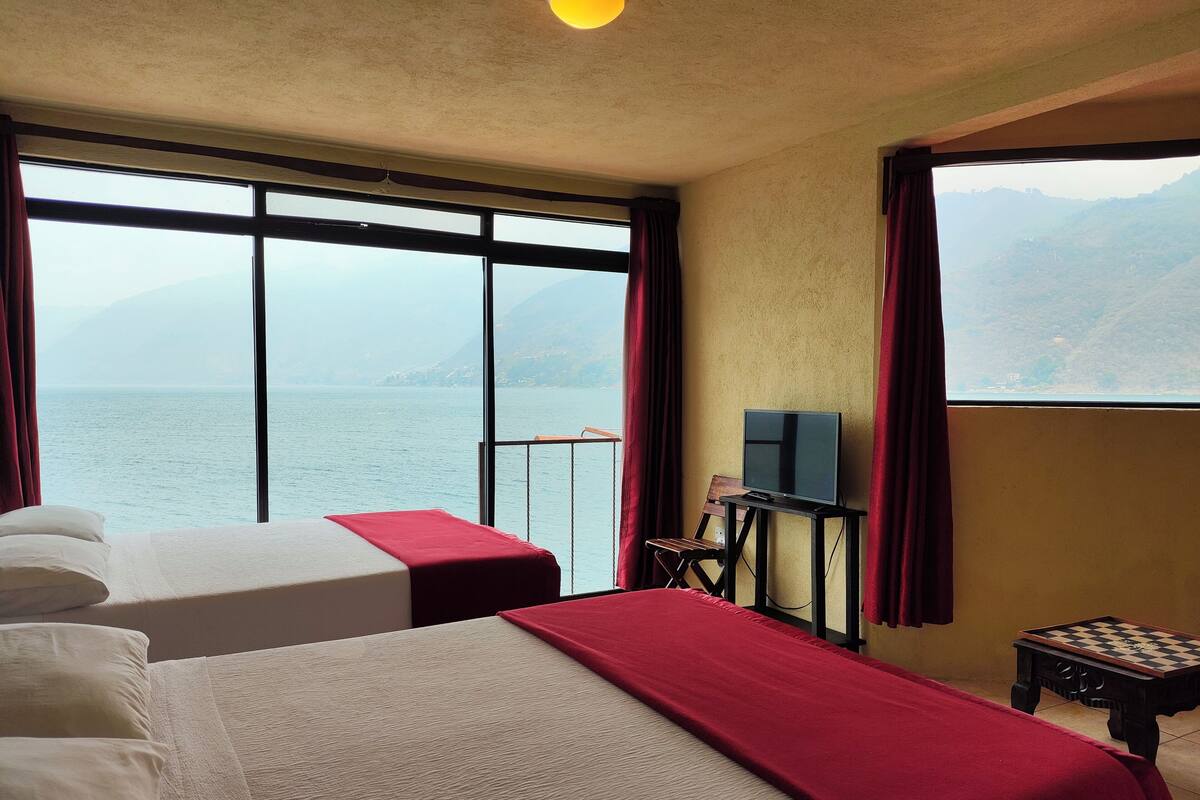 Ein Hotelzimmer mit zwei Betten, einem Fernseher auf einem Schrank, einem kleinen Tisch und Blick auf das Meer und die Berge.