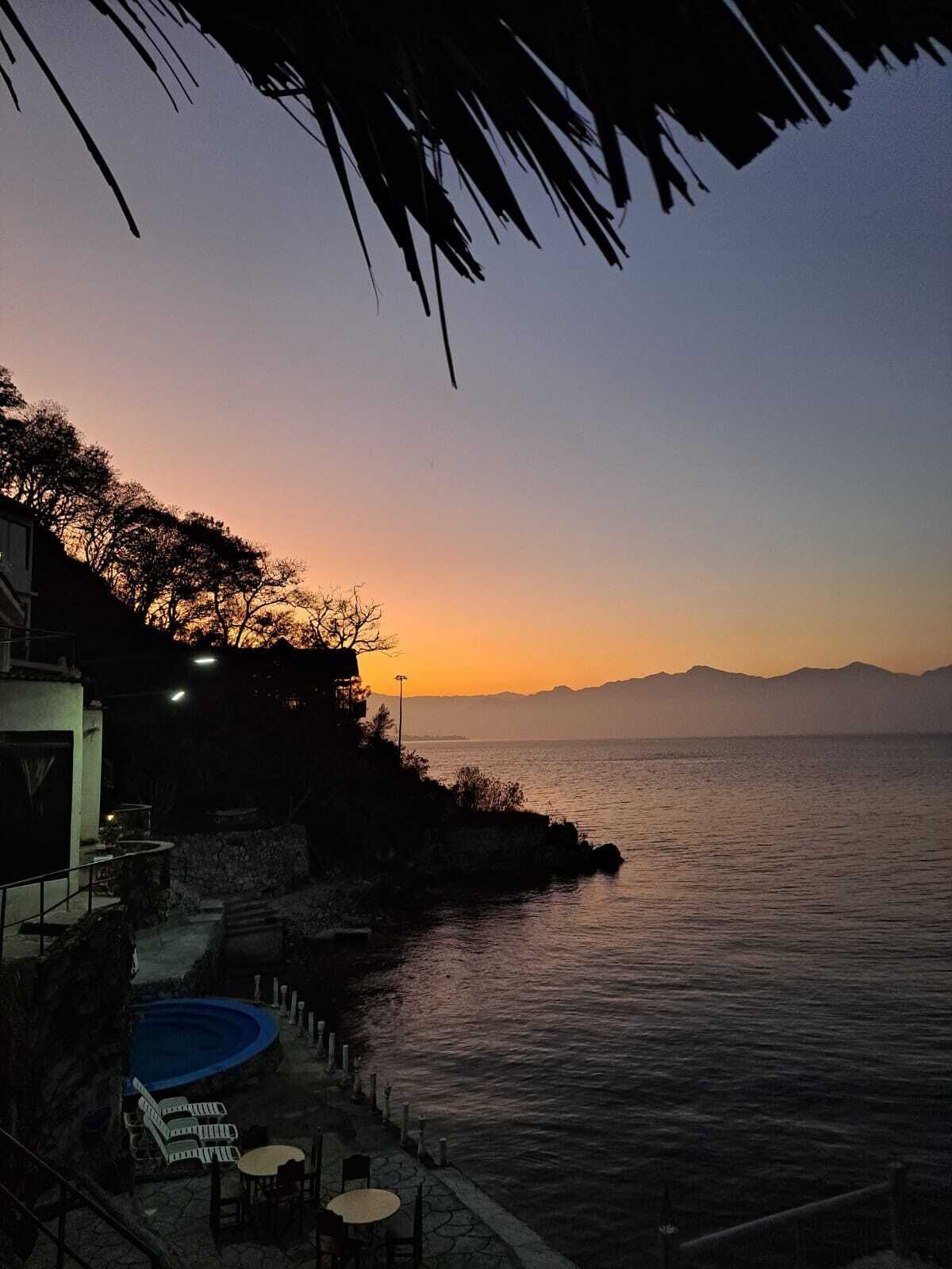 A coastal view with a pool area, outdoor seating, and a sunset over the sea.