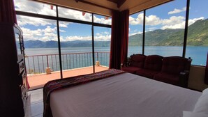 A hotel room with a large bed, a balcony with a red railing, and a view of a lake and mountains. - Hotel La Villa de los Dioses (San Antonio Palopo)