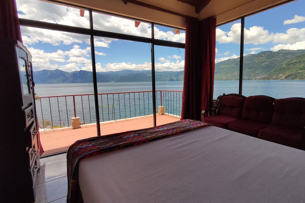 Ein Hotelzimmer mit einem großen Bett, einem Balkon mit roter Brüstung und Blick auf einen See und Berge.