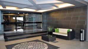 A modern lobby with a glass entrance, a reception desk, a sofa, a coffee table, and a trash can. - Hotel Abastos Plaza (Mexico City)