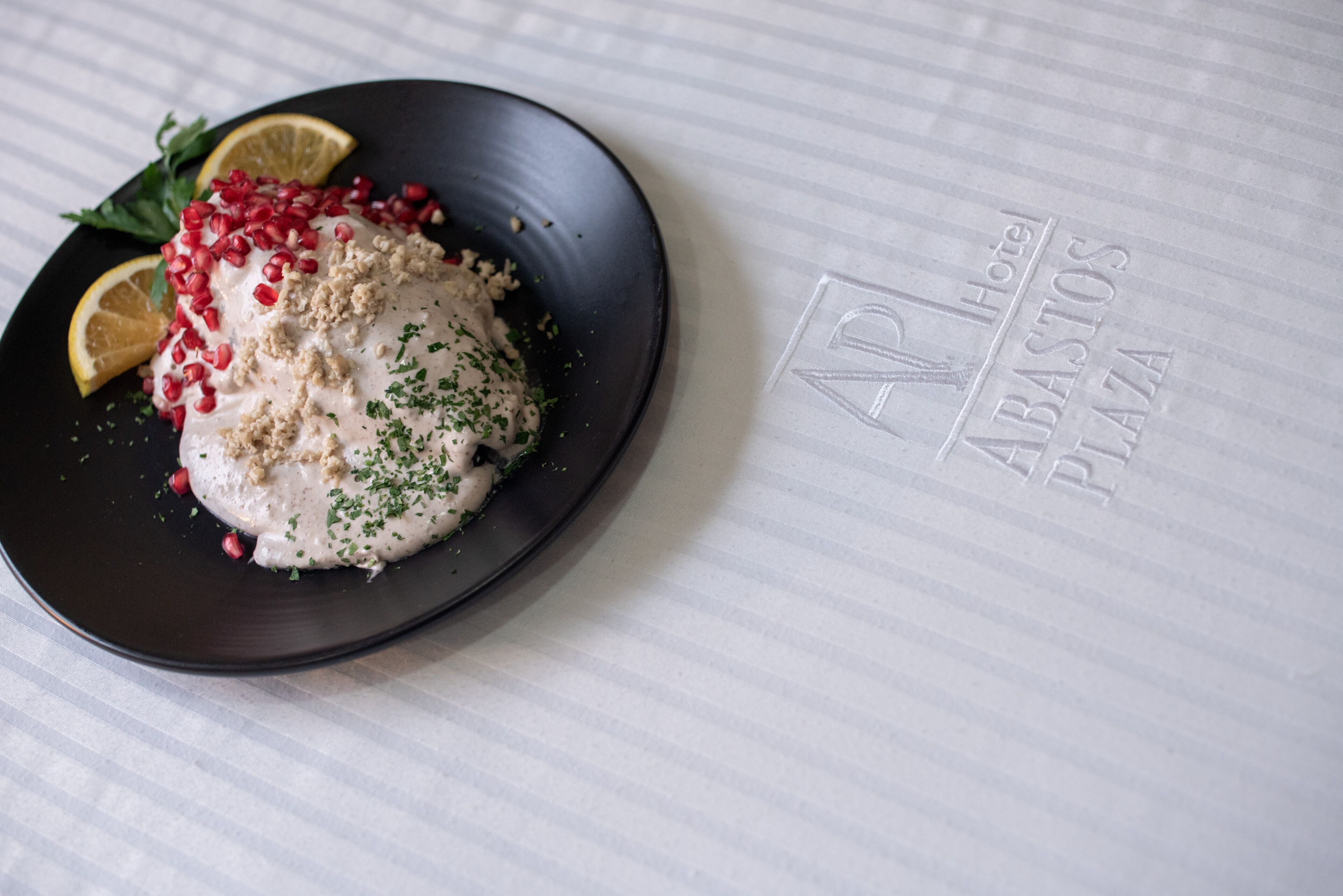 A plate of food with pomegranate seeds and a lemon wedge on a black plate, placed on a table with a hotel logo.
