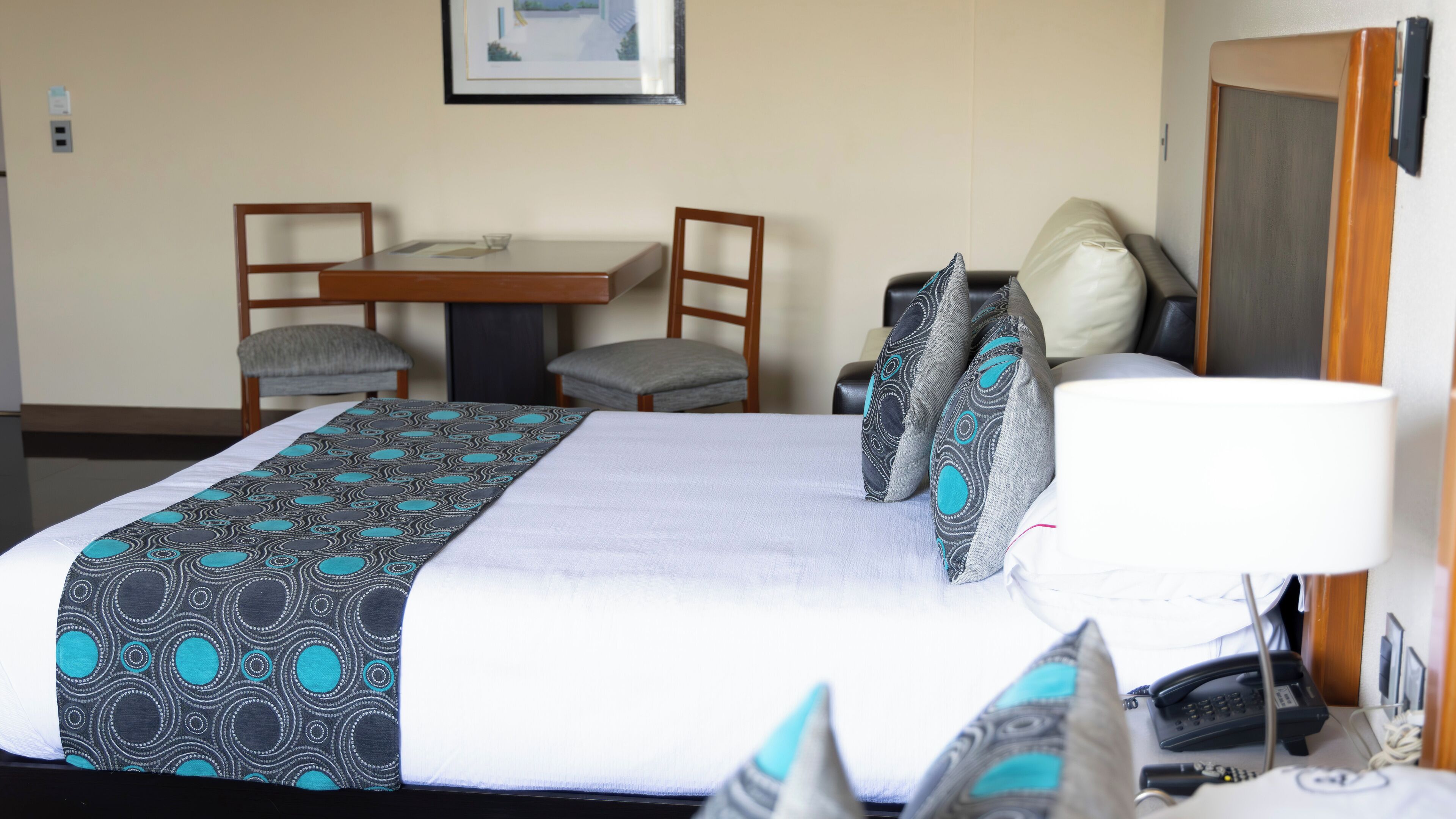 A hotel room with a bed, a desk, two chairs, a telephone, and a wall-mounted picture.