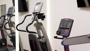 Una bicicleta estática con pantalla digital y panel de control, un televisor montado en la pared, y una puerta al fondo.