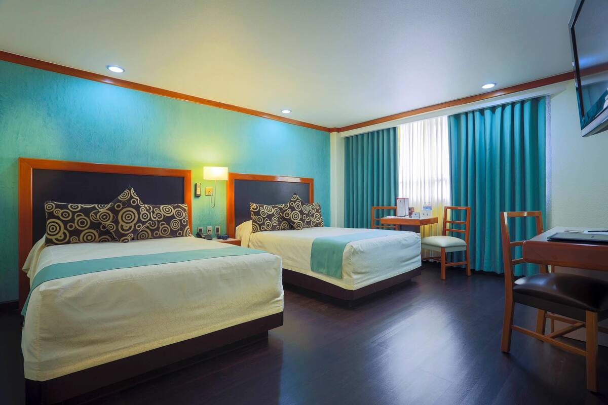 A hotel room with two beds, a desk, a chair, a TV, and a window with curtains.
