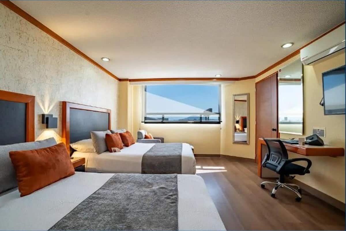 A hotel room with two beds, a desk, a chair, a large window with a view, and a mirror.
