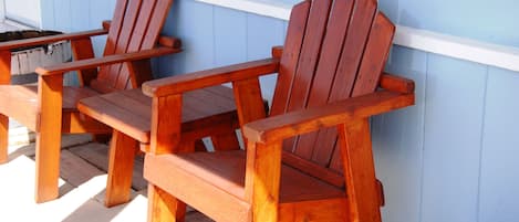 Zwei hölzerne Adirondack-Stühle und eine weiße Holzbank stehen auf einer Holzterrasse vor einer hellblauen Wand.