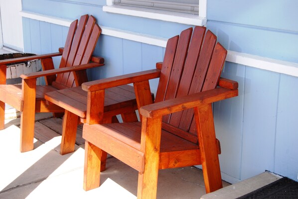 Two wooden Adirondack chairs with a white wooden bench, positioned on a wooden deck against a light blue wall.