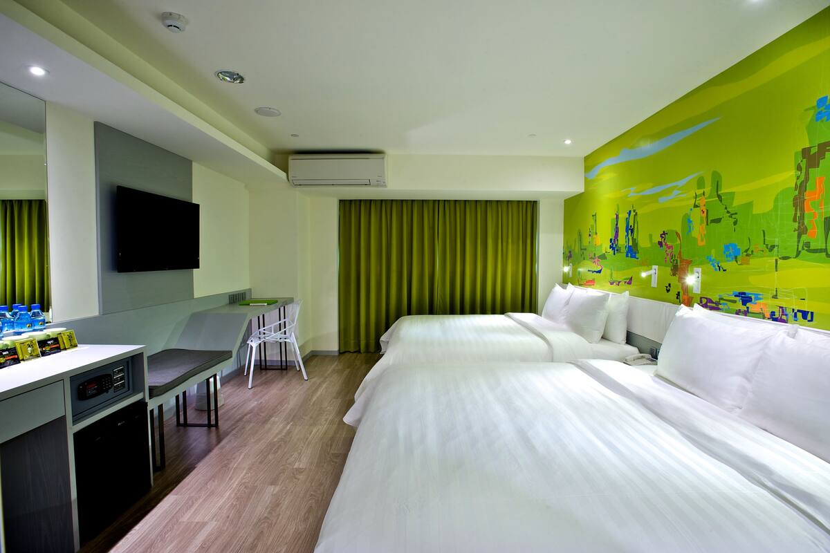 Ein Hotelzimmer mit zwei Betten, einem Schreibtisch mit Stuhl, einem Flachbildfernseher und einem grünen Vorhang.