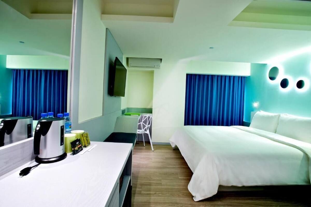 Ein Hotelzimmer mit einem Bett, einem Schreibtisch mit Kaffeemaschine, einem Sessel und einem Fernseher an der Wand.