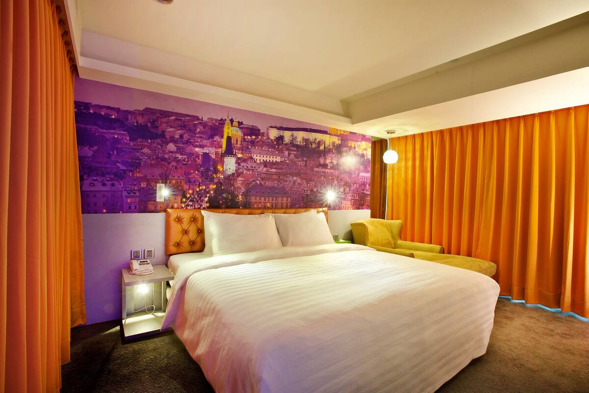 Ein Hotelzimmer mit einem großen Bett, einem Nachttisch mit Telefon, Wandgemälde und orangefarbenen Vorhängen.