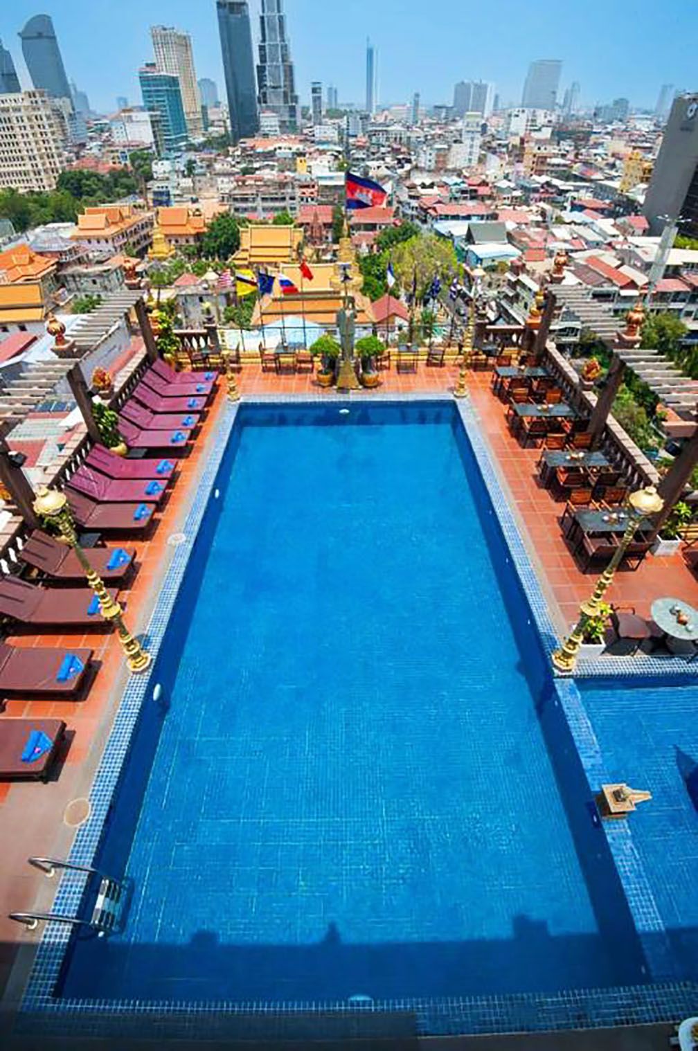 A rectangular swimming pool with a tiled edge, surrounded by lounge chairs and tables, set against a cityscape with buildings and flags.