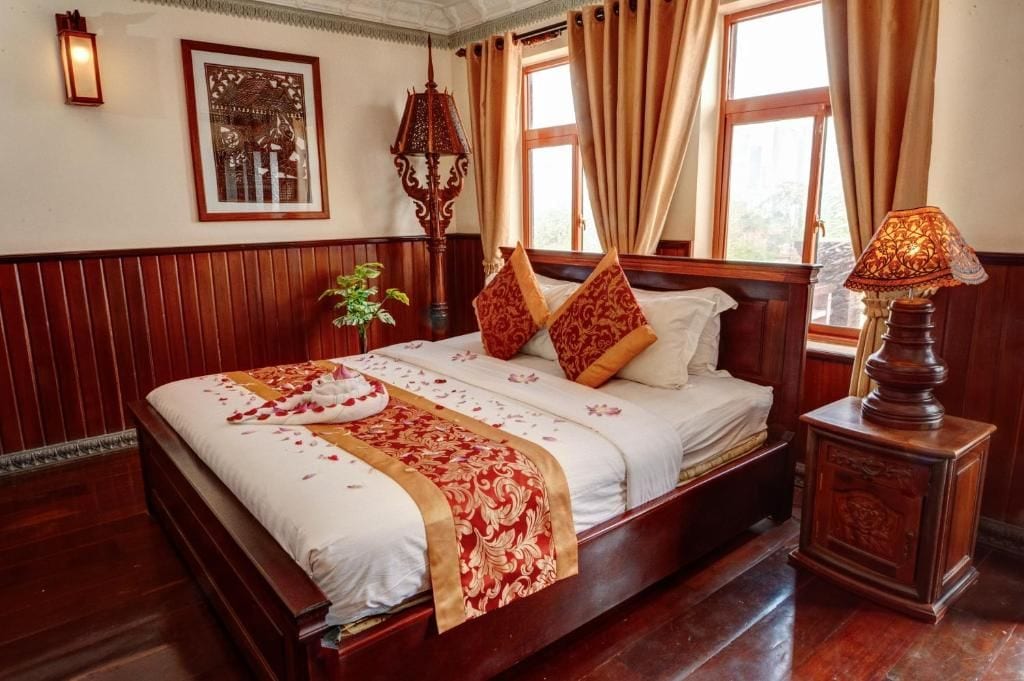 Habitación de hotel con una cama grande, una mesita de noche con lámpara, una planta en maceta y una ventana con cortinas.