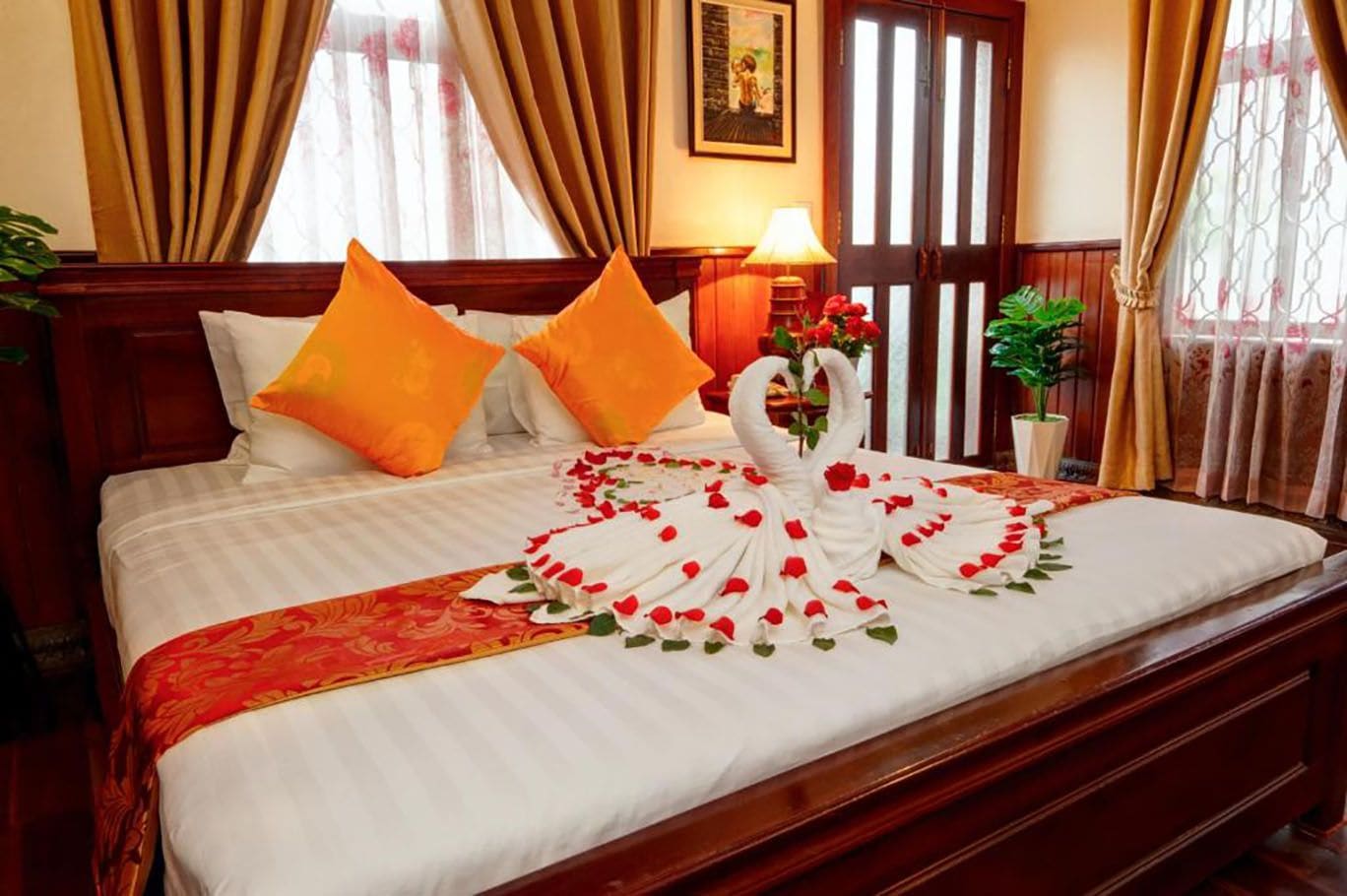 Habitación de hotel con cama, dos almohadas, un cisne decorativo hecho de pétalos de rosa y una colcha con estampado rojo y dorado.