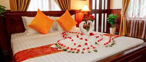 Habitación de hotel con cama, dos almohadas, un cisne decorativo hecho de pétalos de rosa y una colcha con estampado rojo y dorado.