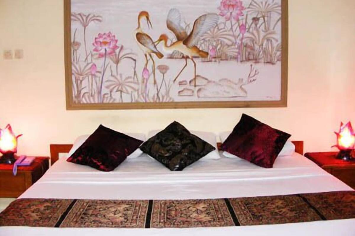 A hotel room with a large painting of birds and flowers on the wall, a bed with a white bedspread and two decorative pillows, and a patterned rug on the floor.