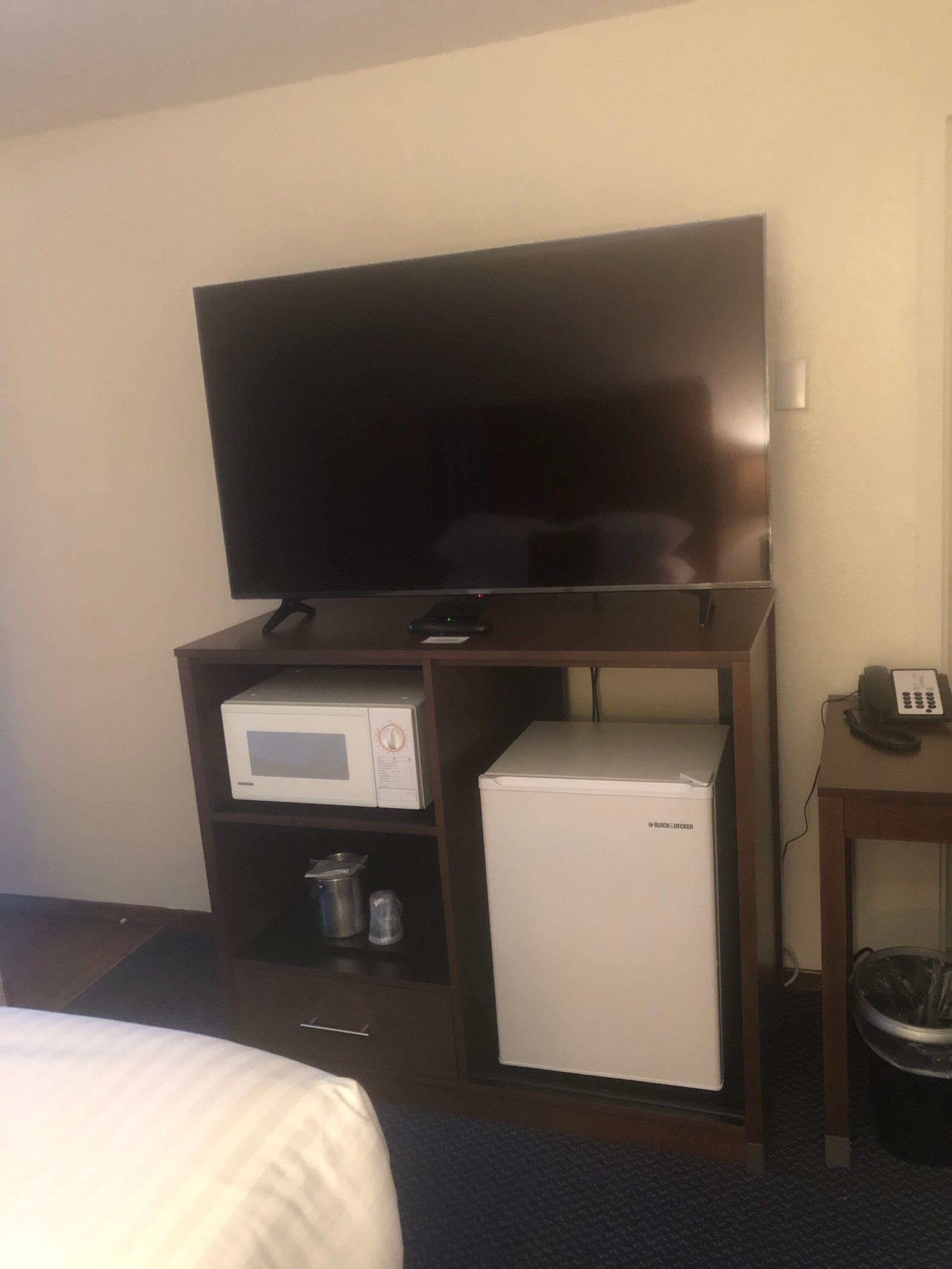 Habitación de hotel con televisión de pantalla plana sobre un soporte de madera, microondas, mini refrigerador, un pequeño cesto de basura y un teléfono sobre una mesita de noche.