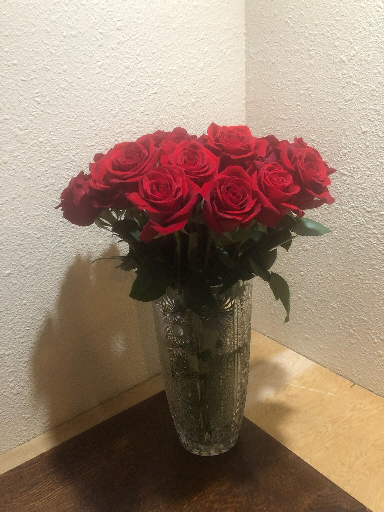 A clear glass vase with a floral arrangement of red roses on a wooden table against a textured wall.