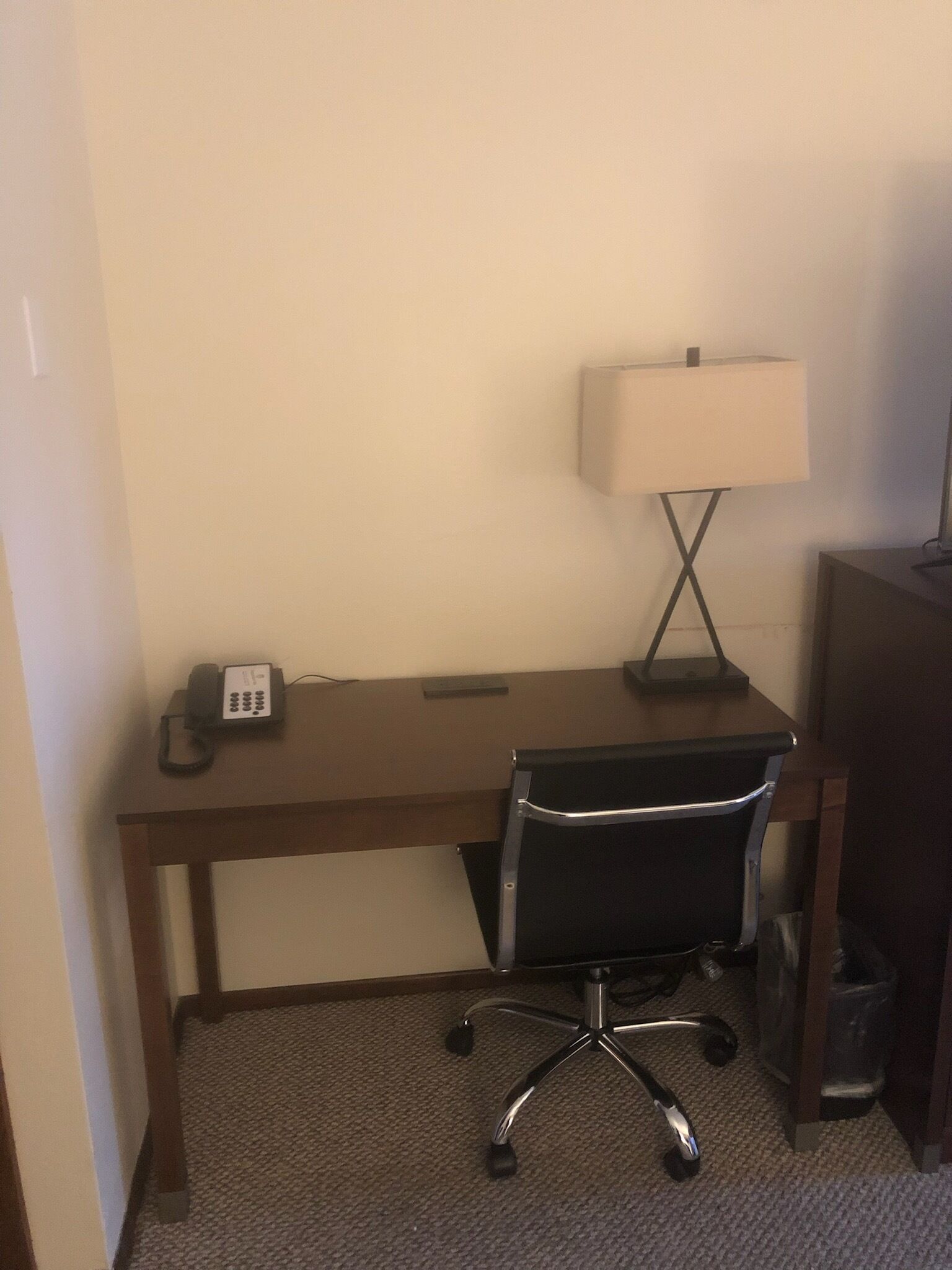 Quarto de hotel com escrivaninha de madeira, cadeira de escritório preta, luminária de mesa e telefone.