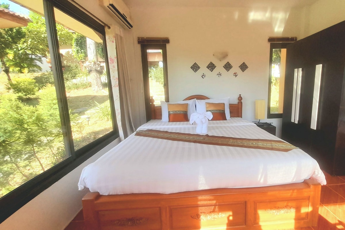 A bedroom with a large bed, a wooden headboard, a nightstand, a TV, and a view of greenery outside.