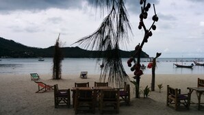 Un café frente a la playa con mesas y sillas de madera, una palmera y barcos al fondo.