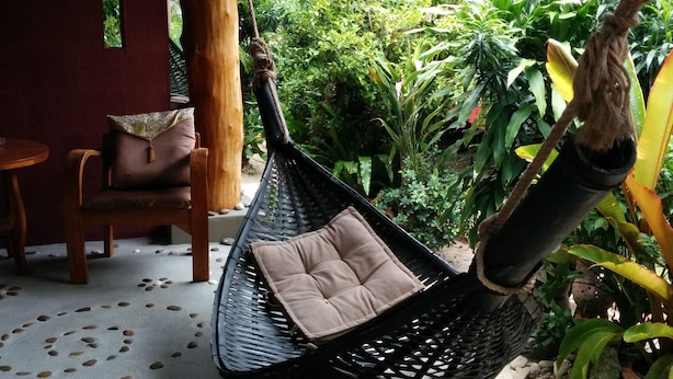 Una silla hamaca con cojines, una mesa de madera y sillas en un espacio exterior cubierto con vistas a exuberante vegetación.