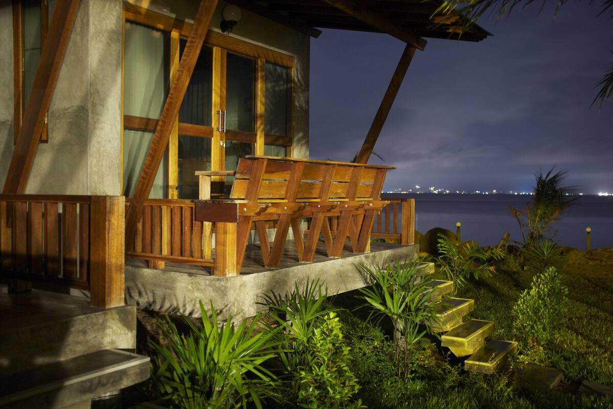 Una terraza de madera con una silla, escalones que llevan a un edificio y vistas del agua de noche.