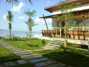 A modern building with a stone pathway leading to a wooden deck, surrounded by greenery and overlooking a body of water. - Phangan Palm Beach Resort (Ko Pha-ngan)