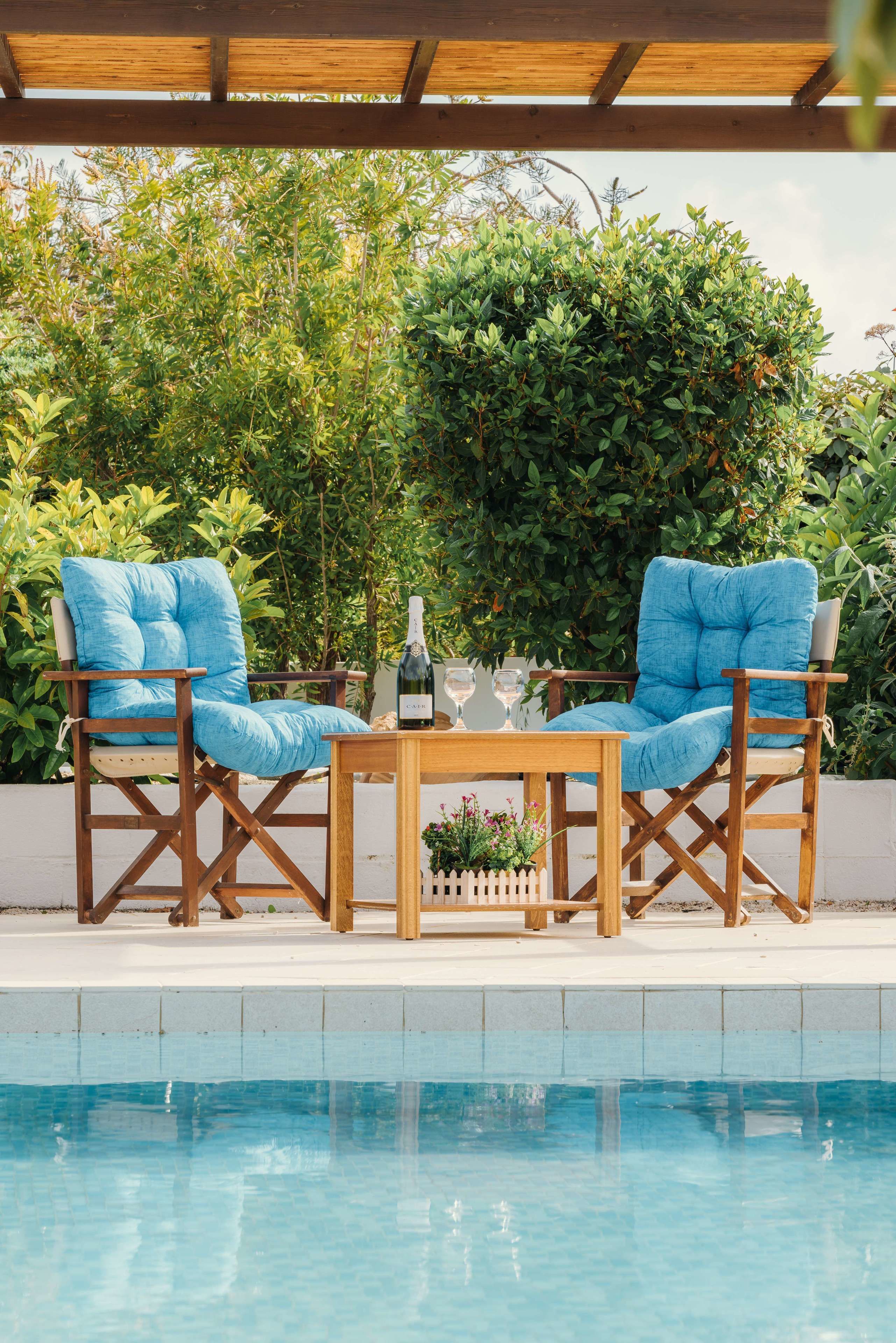 Un área de estar al aire libre con dos sillones azules acolchados y una mesa de madera, con abundante follaje y una piscina de fondo.