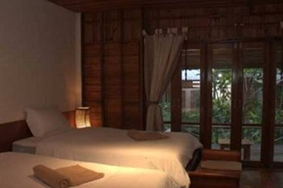 Una habitación con dos camas, una mesita de noche, una lámpara y vistas al exterior con vegetación.