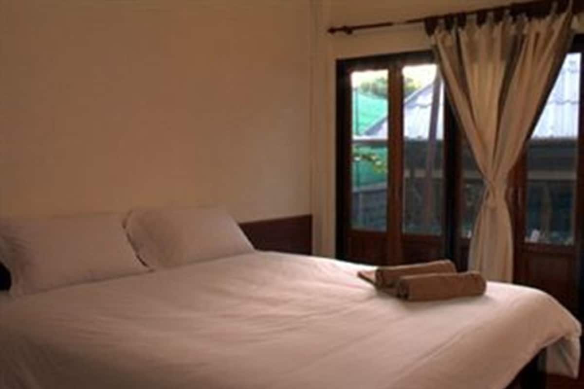 Una cama bien hecha con una almohada y una toalla doblada, una ventana con cortinas y vistas al exterior.