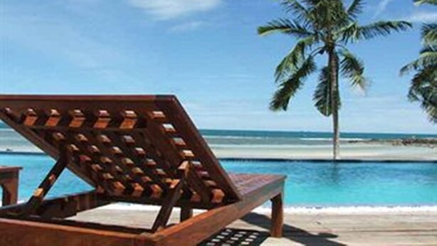 A wooden lounge chair with a metal frame, positioned on a sandy beach with clear blue water and palm trees in the background. - Sunset Cove Resort (Ko Pha-ngan)