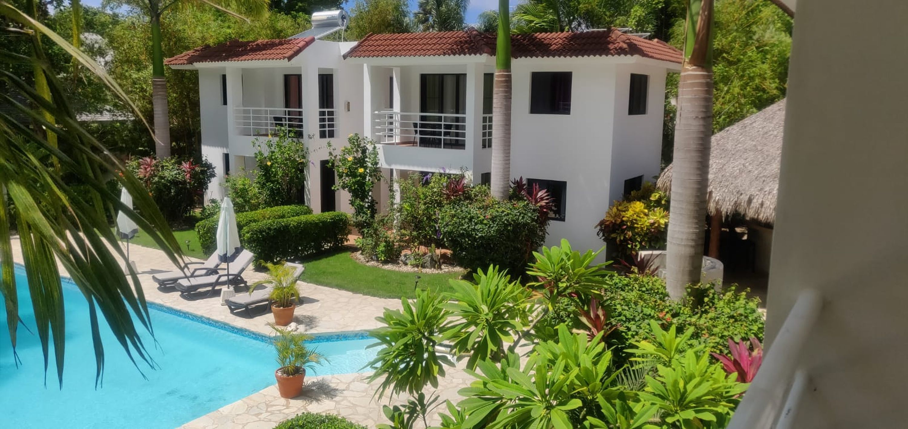 A two-story white building with a red-tiled roof, surrounded by lush greenery and a swimming pool.