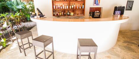 Una zona de bar con mostrador de madera, taburetes de mimbre y estantes llenos de botellas de licores.