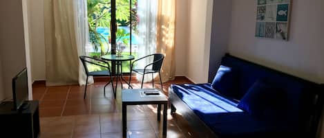 A room with a blue sofa, a small table, two chairs, a TV, and a window with curtains.