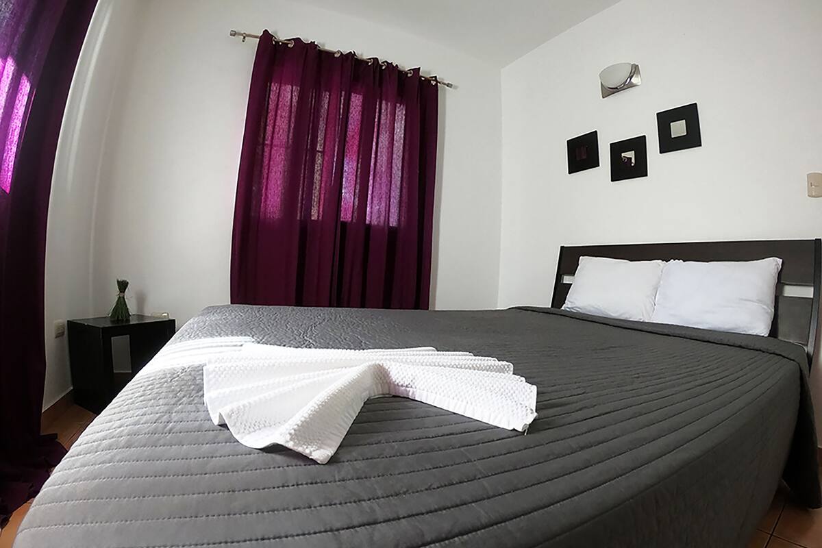 Una cama bien hecha con una colcha blanca, un cabecero oscuro y una ventana con cortinas moradas.