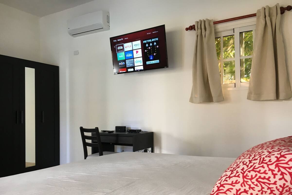 Habitación de hotel con cama, escritorio con silla, televisor de pantalla plana montado en la pared y ventana con cortinas.