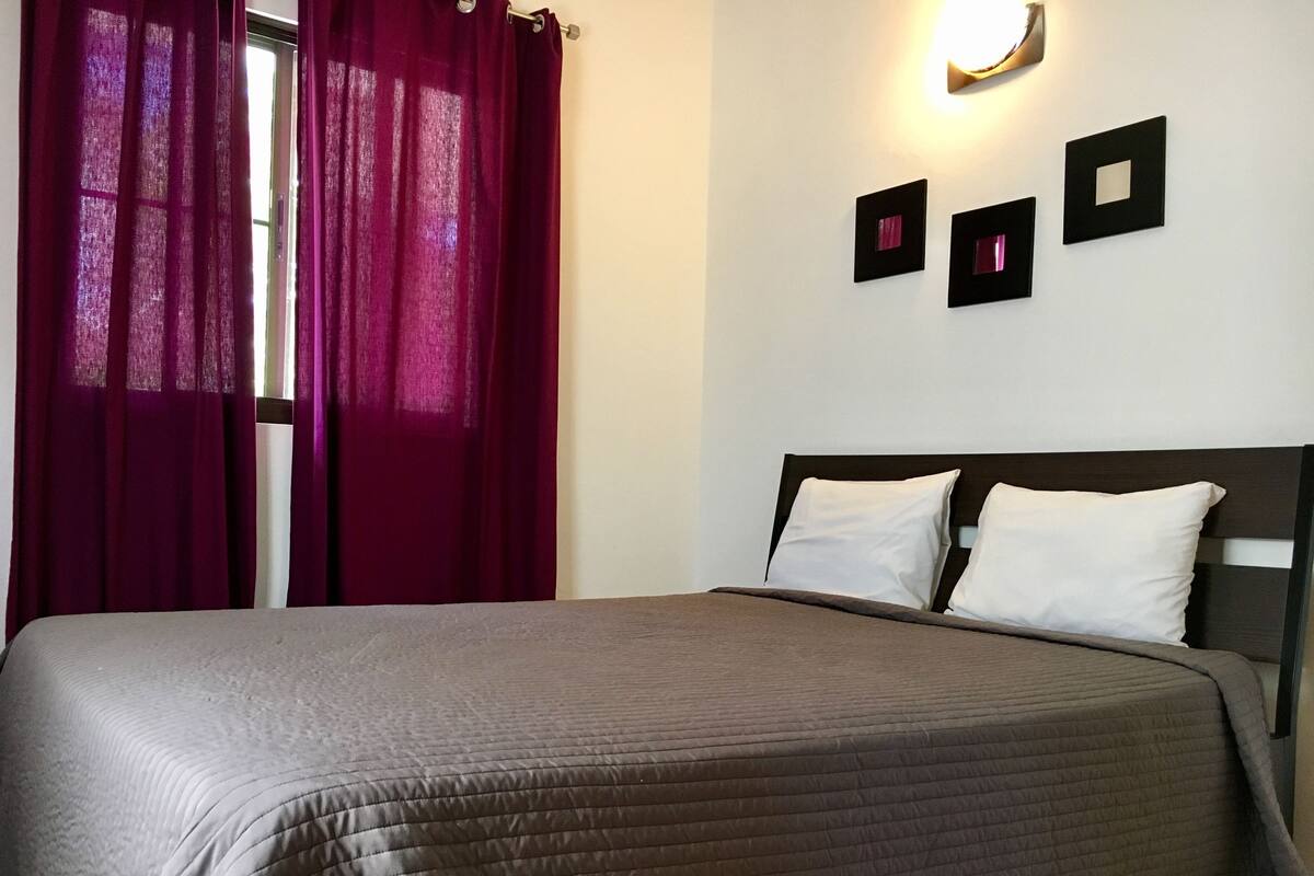 Una cama bien hecha con una colcha oscura, una ventana con cortinas rojas y tres cuadros en la pared.