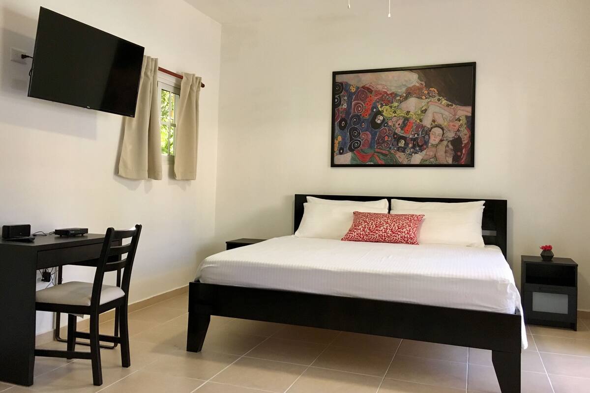 Un dormitorio con cama, escritorio, silla, televisor montado en la pared y un cuadro.